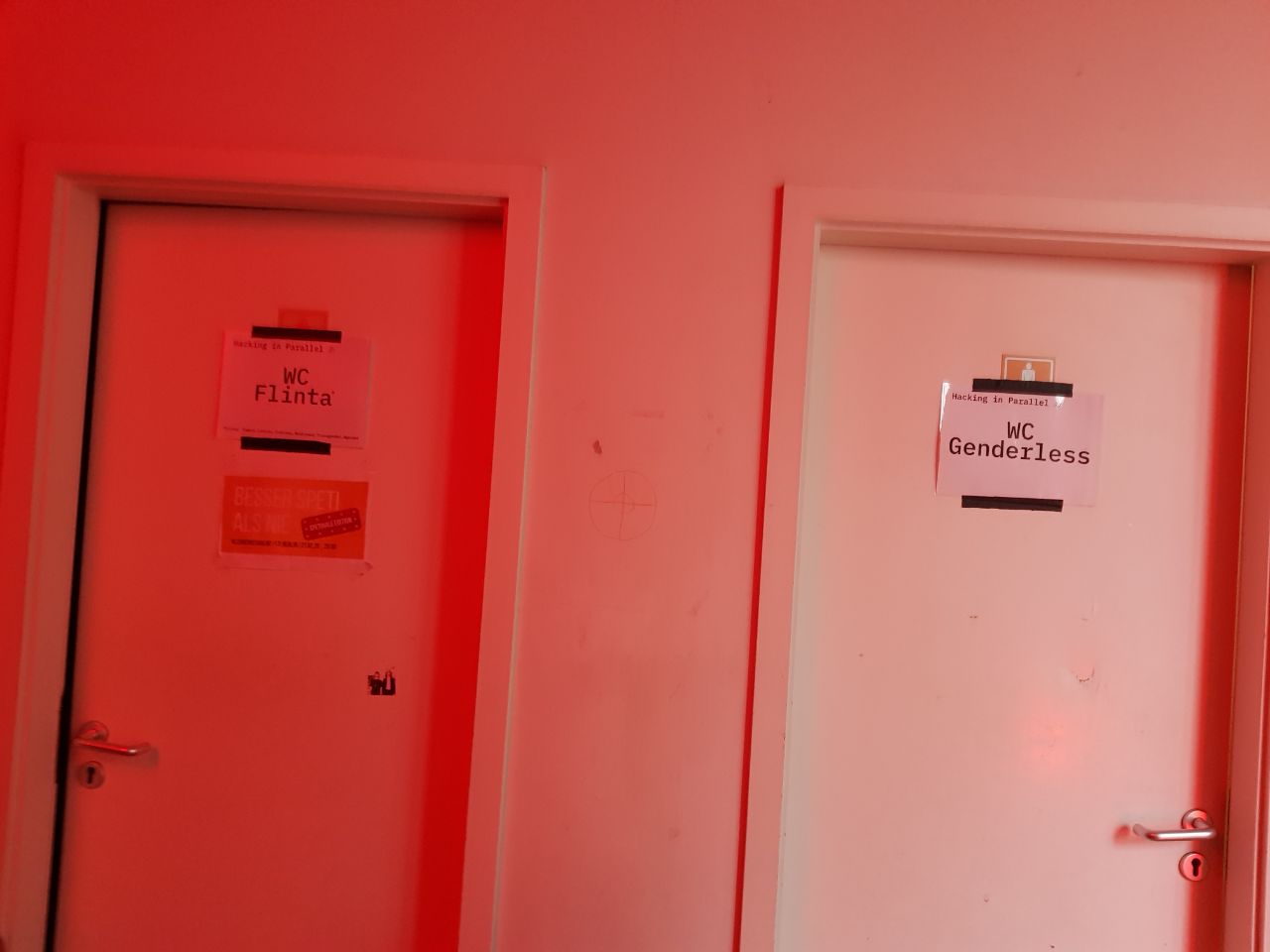 Two toilet doors next to each other, both labeled with pieces of paper hiding the conventional male/female symbols, left piece of paper says "WC FLINTA*", right one says "WC Genderless" (and both peices of paper having printed "Hacking in Parallel" in much smaller letters on top).